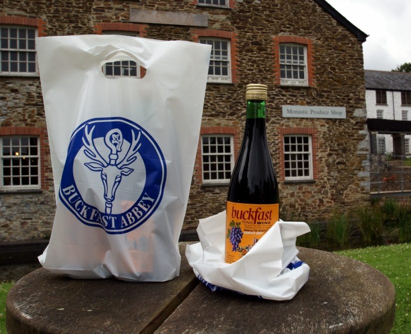 A photo of a back labeled "Buckfast Abbey" next to a bottle labeled "Buckfast" in front of a stone building with the sign "Monastic Produce Shop" above one window
