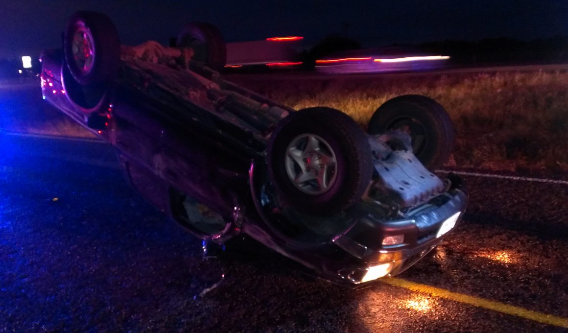 Photo of an overturned vehicle on the shoulder of a road at night, headlights still shining