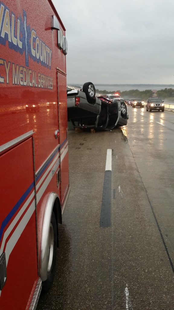 Side of ambulance and overturned automobile on a highway, wet with rain, under an overcast sky, with approaching cars, headlights shining through the mist