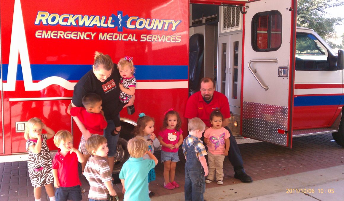Image of ambulance parked next to sidewalk, with two adults and ten children.
